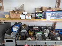 Shelf with grey organiser bins filled with brass door hardware, wire spools, cables, conduit, clear storage boxes and Schlage box