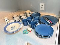 Whole lot on countertop: plates, mugs, bowls, knives, salt & pepper, pitcher
