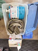 Top view of open plastic tote showing sleeping bag and water jug; boxed toilet, blue bag and cleaner bottle alongside