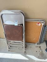 Stack of three beige metal folding chairs with brown vinyl seats/backs leaning against folded card table
