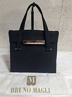Front view of black canvas handbag with metal Bruno Magli logo plaque and accompanying dust bag