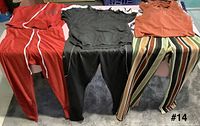 Five visible garments laid out on bed: red track pants, black T-shirt, black jogger pants, orange bodysuit, multicolour striped pants