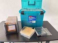 Overall lot showing teal Rubbermaid Box Office bin filled with CD cases, black stackable holder, and stacks of cases