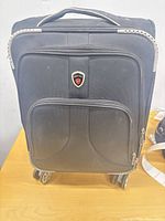 Front view of black Goodpartner carry-on suitcase showing logo badge, exterior pocket, wheels, and top handle