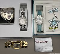 All items laid out: two boxed Gruen watches, gold-tone square-link watch, two rhinestone brooches, heart-shaped accessory, Gruen booklet