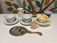 Group shot of all items: three teacup sets, mug, planter, miniature cup, brass pelican, brass hand mirror