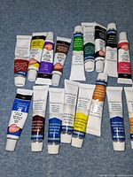 Array of 16 acrylic paint tubes laid out on blue surface