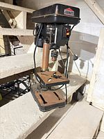 Full view of Jobmate bench drill press on stair tread with accessories on table