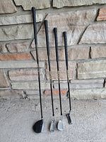 Four junior golf clubs standing upright against stone wall: driver/wood, 5 iron, 9 iron, putter
