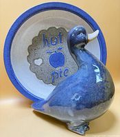 Front view of blue glazed duck figurine positioned before interior of bowl showing "hot pie" design.