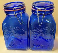 Front view of both cobalt blue glass canisters showing embossed fruit design and metal bail lids