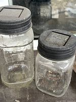 Front view of both jars showing size difference, clear glass bodies, black lids with square recess, copper wire visible in smaller jar