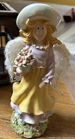 Front view of pastel ceramic angel figurine with yellow skirt, lavender blouse, white hat, lace wings, holding pot of pink flowers