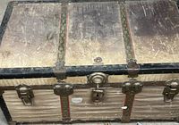 Top and front view of tan steamer trunk showing black trim, red stripes, brass hardware, wear