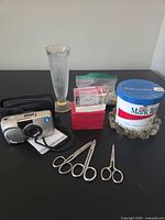 Overview of all items on tabletop: camera with case, scissors, flute glass, red box with acrylic blocks, puzzle pieces bag, tobacco container set in glass ashtray