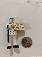 Colonel Sanders figurine beside coin showing 25 Years side