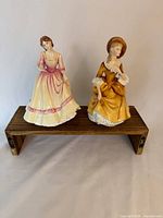 Both Royal Doulton figurines side by side on small wooden riser