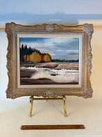 Full view of framed oil landscape painting on easel with ruler for scale