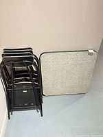 Stacked four black metal folding chairs and folded square table front with gray laminate top