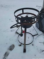 Full view of propane burner stand on snow showing circular pot ring, legs, hose, regulator