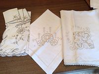 Three folded tea cloths and folded napkins showing cutwork, embroidery and edges