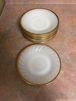 Stack of ten white milk glass saucers with gold rims displayed on countertop