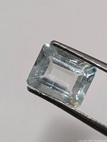 rectangular emerald-cut aquamarine held by tweezers, showing light blue color and clarity