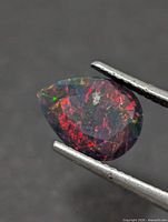 Pear-cut opal gemstone held in tweezers, showing red and green play-of-color