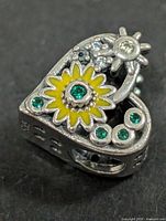 Front view of heart-shaped silver bead with yellow enamel flower and green stones