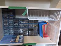 Blue plastic drawer organizers filled with assorted hardware, multicolour stackable bins and clear lid tray on shelf