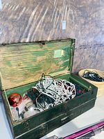 Open green trunk holding tangled white and green wired string lights with ornaments visible, basket of additional lights beside