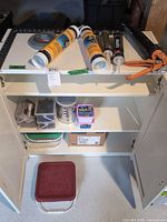 Cabinet showing all items: sealant tubes, caulking gun, tape roll, tubs of screws, box of nails, bucket