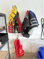 Four life jackets hanging: yellow dog PFD, blue human PFD, black/red human PFD, red dog PFD