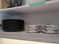 Stack of black stoneware plates beside teacups and saucers on shelf