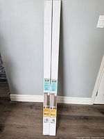 Two unopened long white boxes of blinds leaning against wall