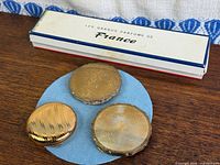 All three gold-tone round compacts grouped on blue pad, decorative box in background