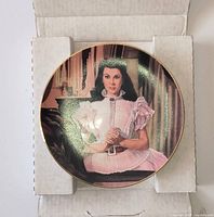 Front view of porcelain collector plate inside open box showing image, gold rim and unused condition