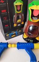 Front view of boxed game beside assembled duck target, blaster and foam balls visible