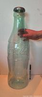 Full view of clear Coca-Cola bottle coin bank standing upright next to pen for scale