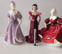 Front view of three English porcelain figurines: lavender standing figure, deep red standing figure, seated red figure