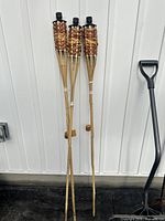 Three bamboo tiki torches leaning against wall next to shovel for scale