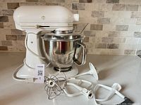 White KitchenAid Artisan stand mixer with stainless bowl and assorted attachments arranged on countertop