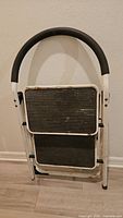 Front view of folded white metal two-step stool with black treads and foam-padded handle