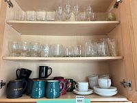 Cabinet shelves holding glassware on two shelves and mugs, Mikasa cups and saucers on bottom shelf