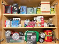 Overview of cabinet shelves containing boxed Noma floodlights, various bulb packs, flashlights and accessories