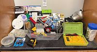 Overall view showing basket of batteries, tools, multimeter in green case, smoke alarm, scissors packaging, tape measure, hardware dishes