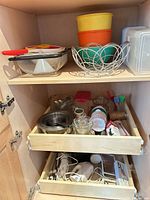 Cabinet showing stainless bowls, glass measuring cups, plastic bowls, strainer, wire basket, food chopper, hand mixer, electric knife, toaster