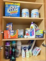 Cabinet shelf showing OxiClean box, two tubs, Bounce dryer sheets, Zout spray, liquid additive jug and other cleaners