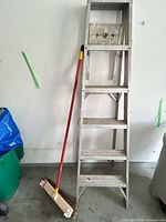 Full view of aluminum ladder standing folded next to broom