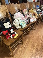 Row of plush toys on chairs including Mickey pillow, small Mickey, pastel bunnies, rag doll and bears